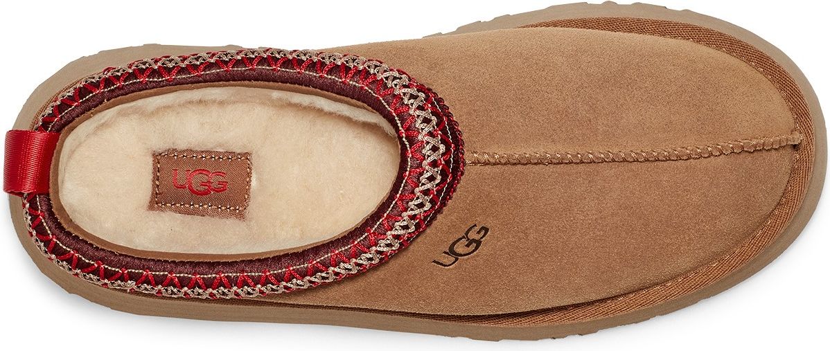A tan-colored slipper featuring a stitched border with red accents around the opening and a red pull tab on the back.